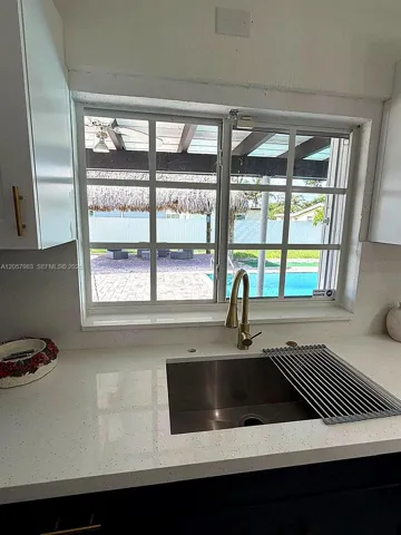White solid surface countertop, undermount stainless steel sink, gold-finish gooseneck faucet, white upper cabinetry with gold-finish pulls, and a window with a grid pattern