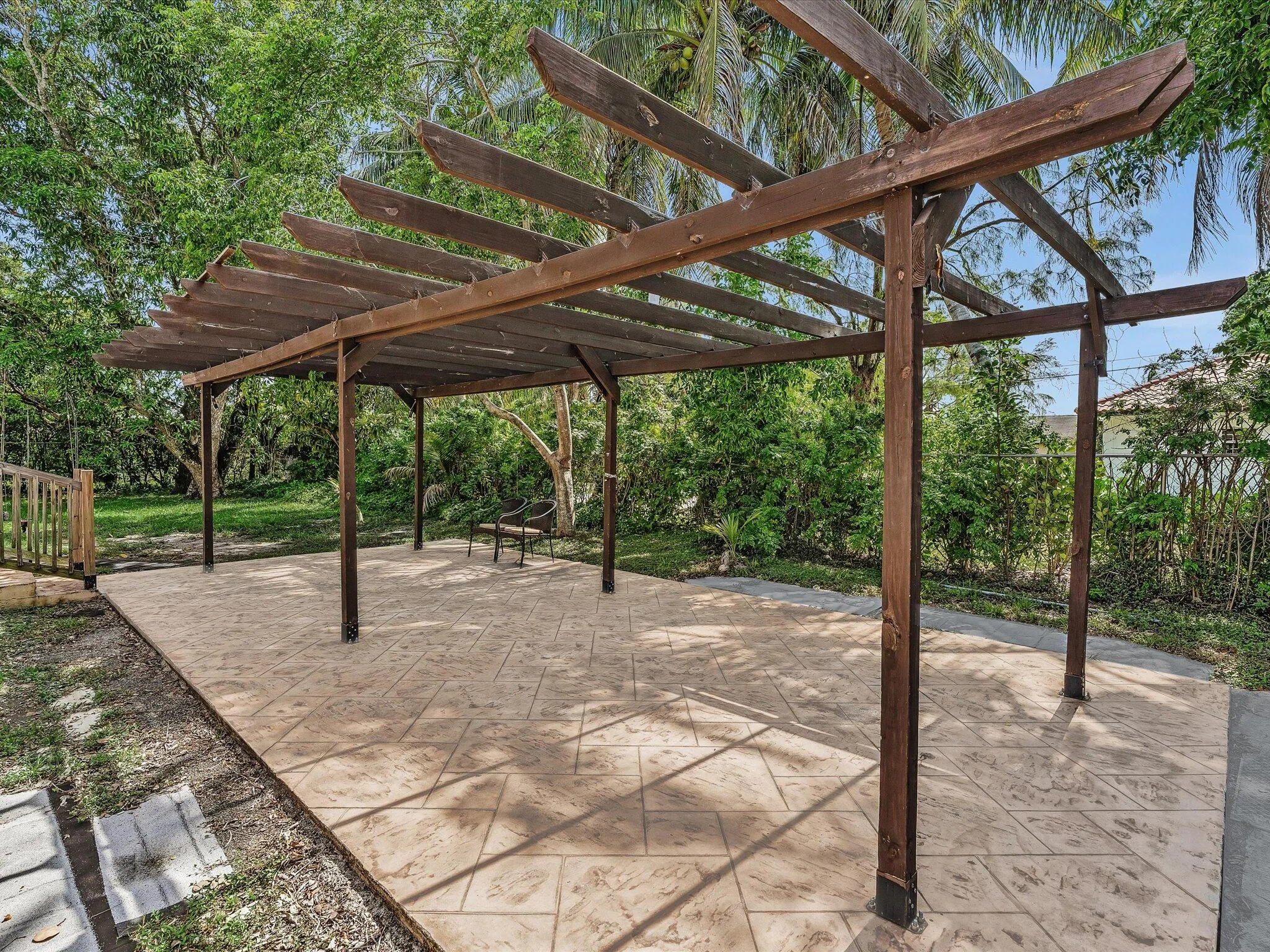 Expansive outdoor patio featuring a large wooden pergola, stamped concrete flooring, lush green landscaping, and mature trees