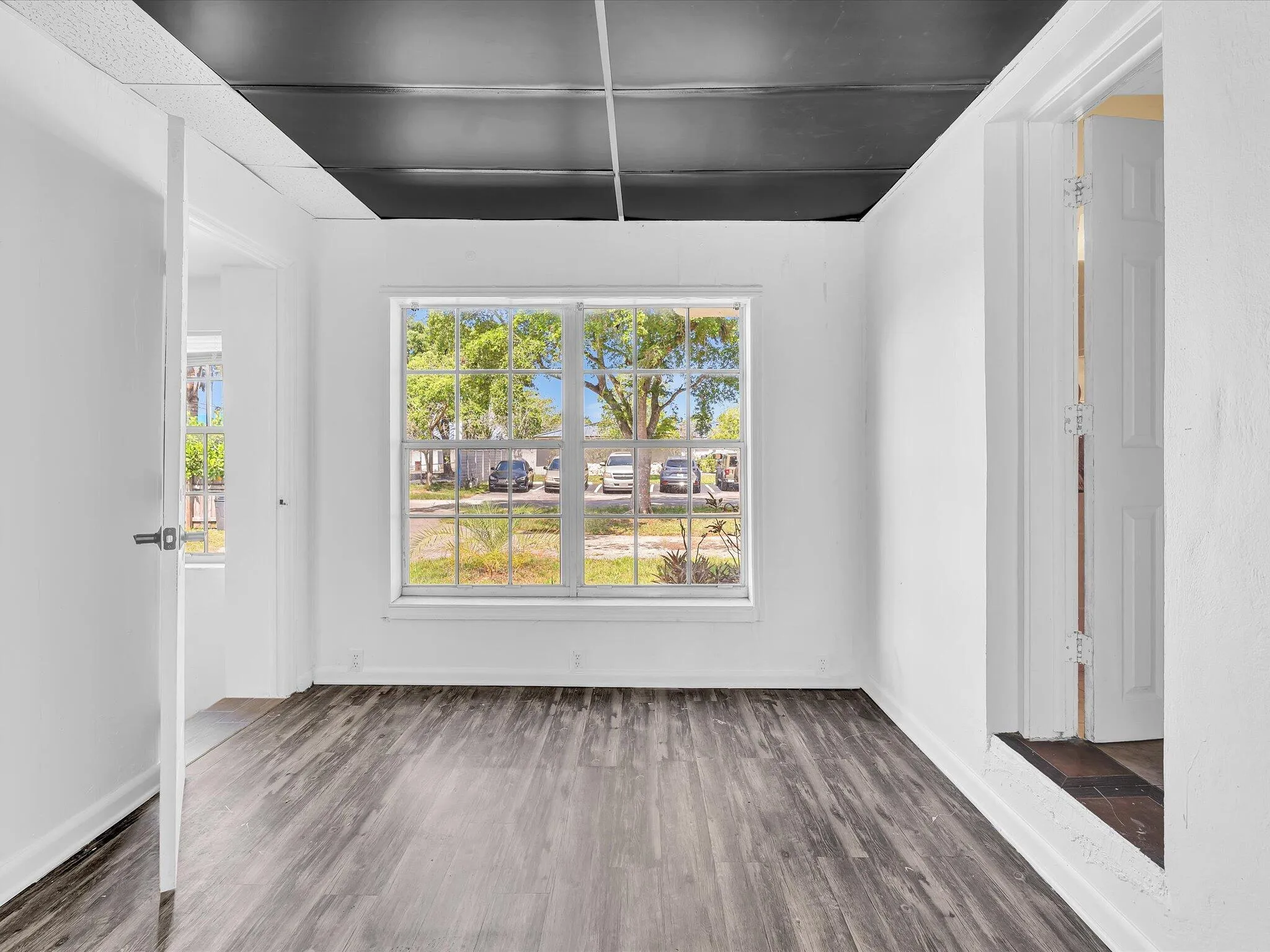 Wood-finish flooring throughout, featuring a large multi-pane window, white painted walls, and a dark ceiling