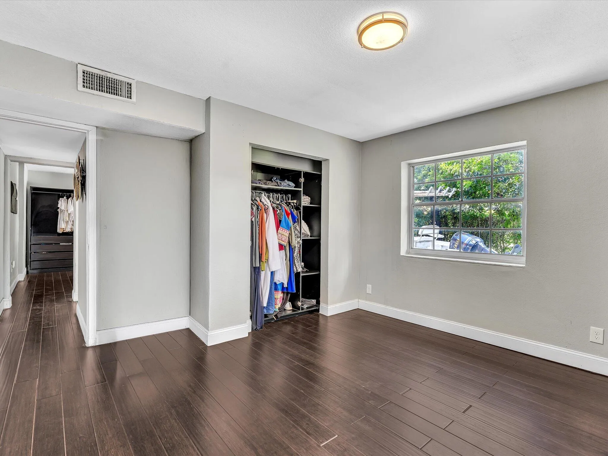 Spacious room featuring rich wood-finish flooring, light gray walls, an integrated closet system with shelving and hanging storage, a rectangular window, and a flush-mount ceiling light fixture