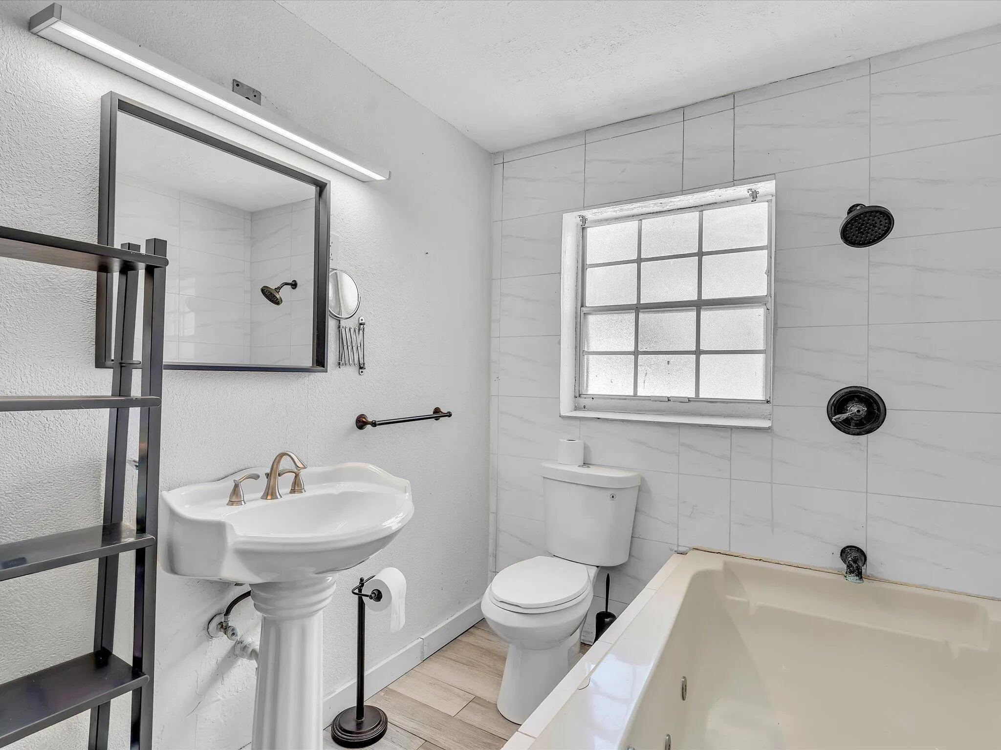 Bathroom featuring a pedestal sink, framed mirror with integrated lighting, tiled walls, a window with grid panes, and a built-in tub with black fixtures