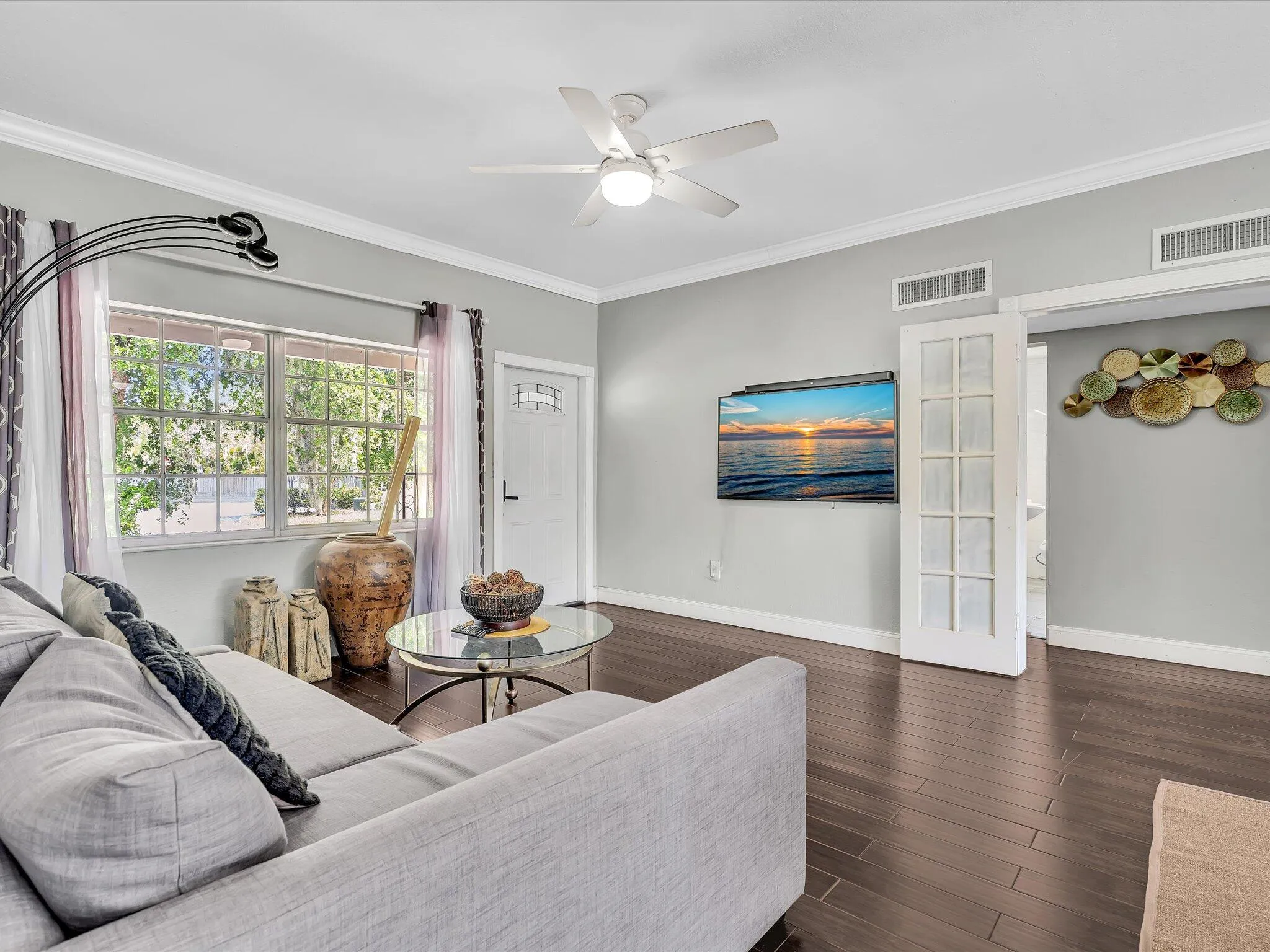 Spacious living area featuring dark wood-finish flooring, ample natural light from multiple windows, crown molding, and a ceiling fan