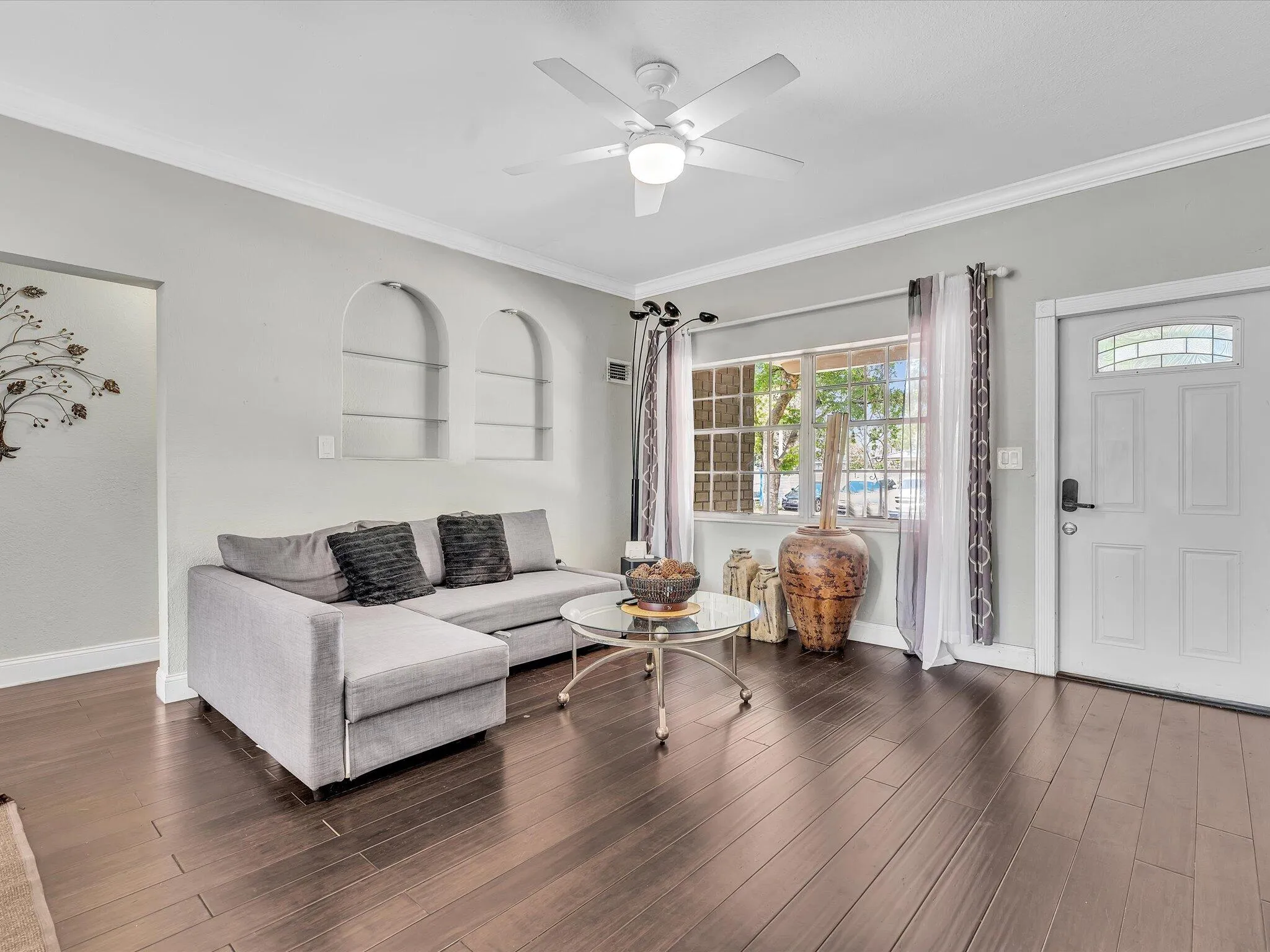 Spacious living area featuring dark wood-finish flooring, a contemporary ceiling fan, dual arched wall niches, a front entry door with decorative glass, and a large window
