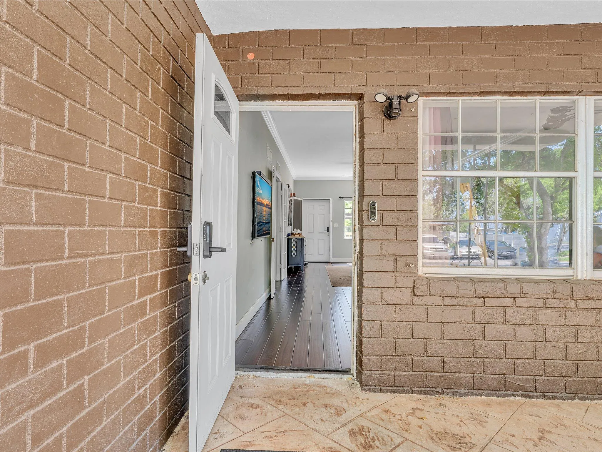 Brick exterior featuring a white entry door with a dark handle, a grid-pattern window, an exterior light fixture, and a tiled patio