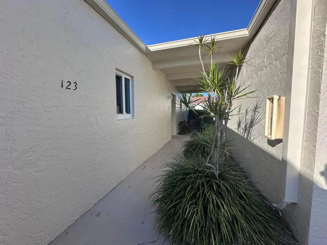 Covered walkway entrance featuring stucco exterior, a single window, and integrated planter beds with mature tropical foliage