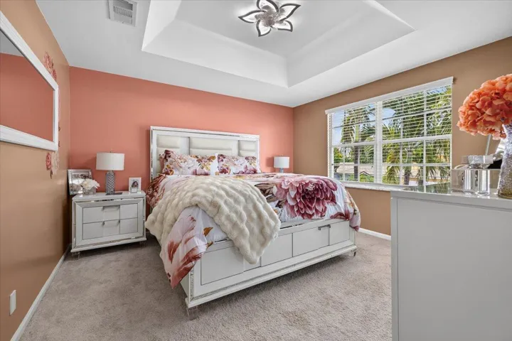 Bedroom featuring a tray ceiling with a decorative light fixture, a large window with blinds, and neutral carpet flooring