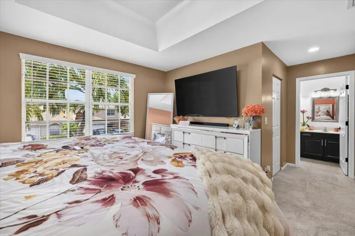 Carpeted room featuring a large window with horizontal blinds, a tray ceiling, and a doorway leading to a bathroom with a dark wood-finish vanity