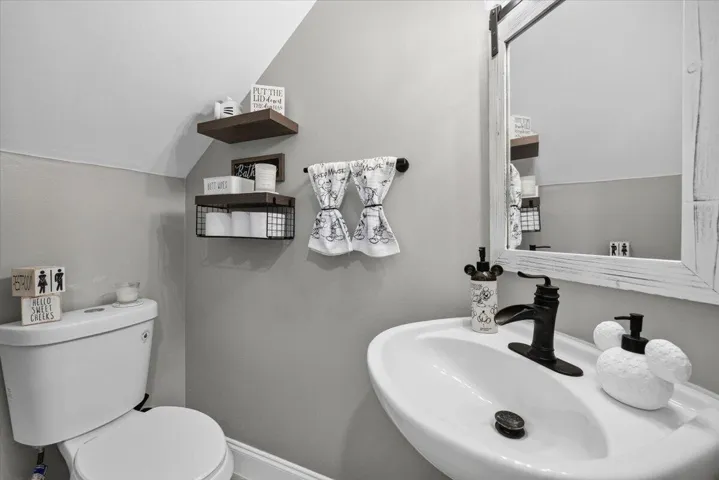 Powder room featuring a pedestal sink, wall-mounted mirror with a distressed white frame, toilet, and floating wooden shelves with metal basket storage