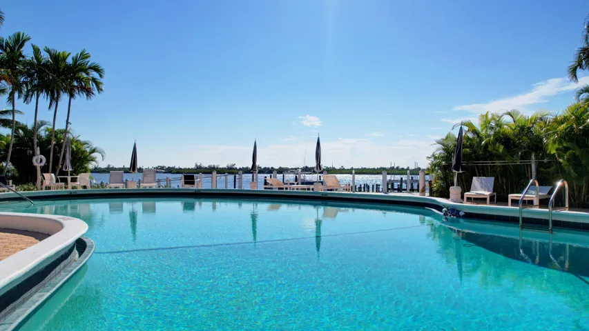 Expansive outdoor swimming pool featuring a brick paver surround, surrounding lush tropical landscaping, and a waterfront setting with a dock structure