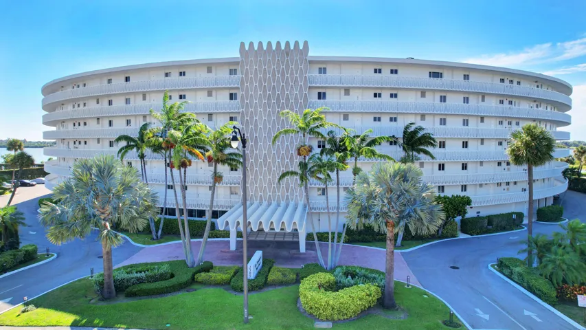 Curved condominium building featuring multiple balconies with white railings, a patterned facade element, a covered main entrance, mature palm trees, and manicured landscaping