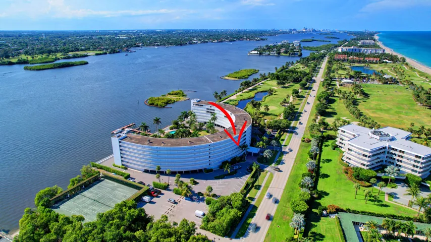 Waterfront residential building featuring a curved facade, multiple balconies, a tennis court, and lush landscaping