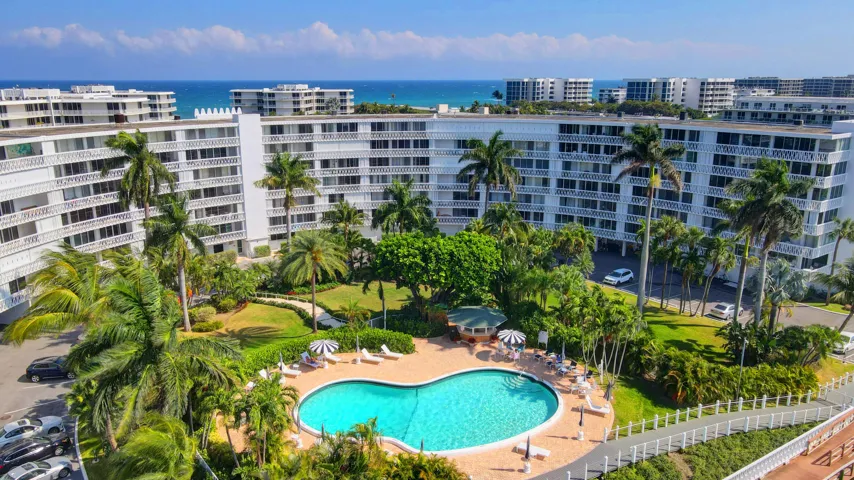 Resort-style residential complex featuring an outdoor swimming pool with surrounding lounge areas, mature landscaping including palm trees, and ocean visible in the background