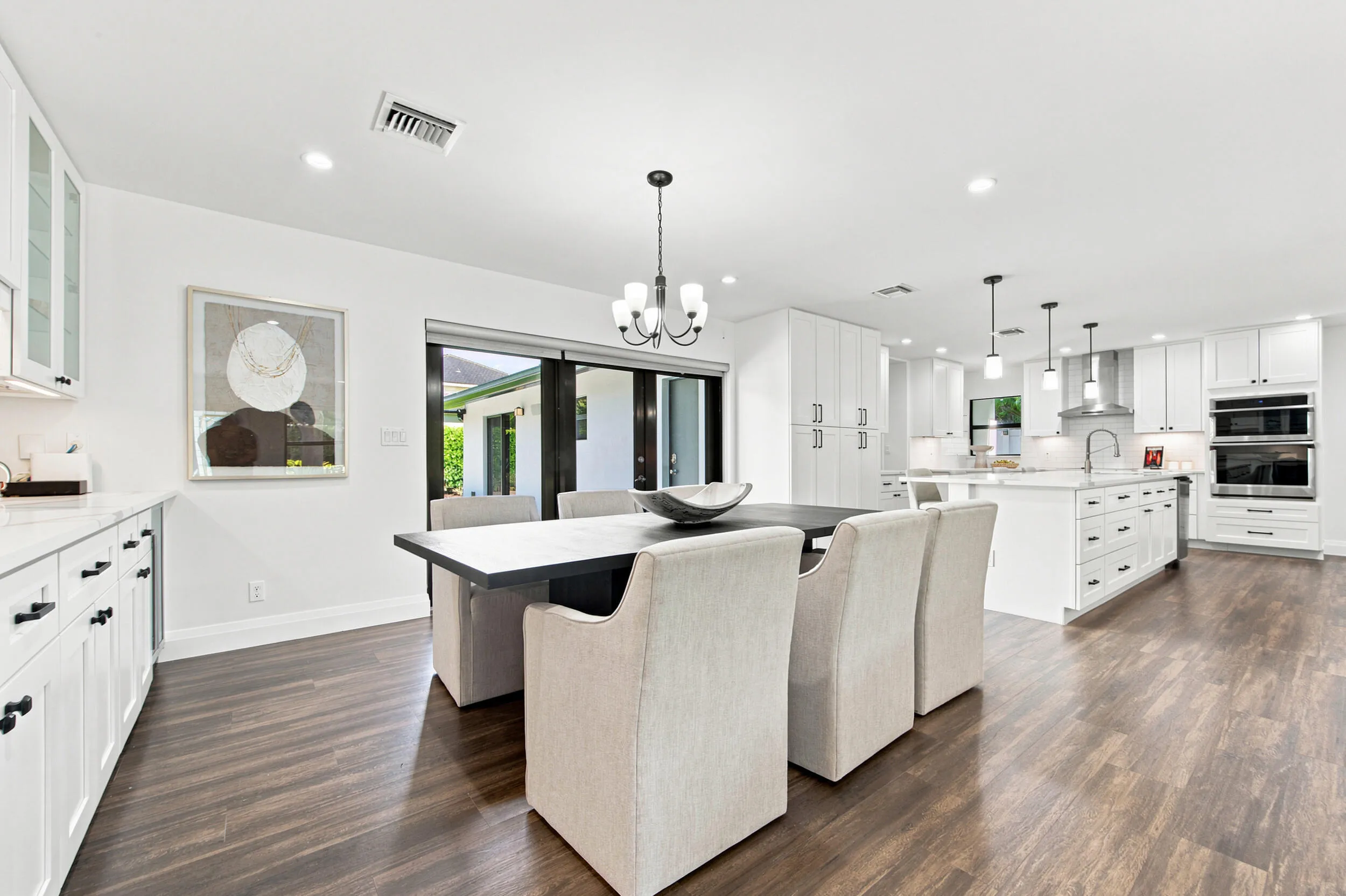 Bright, open-concept kitchen and dining area featuring wood-finish flooring, white cabinetry with dark hardware, a large kitchen island with pendant lighting, stainless steel double wall ovens, and sliding glass doors
