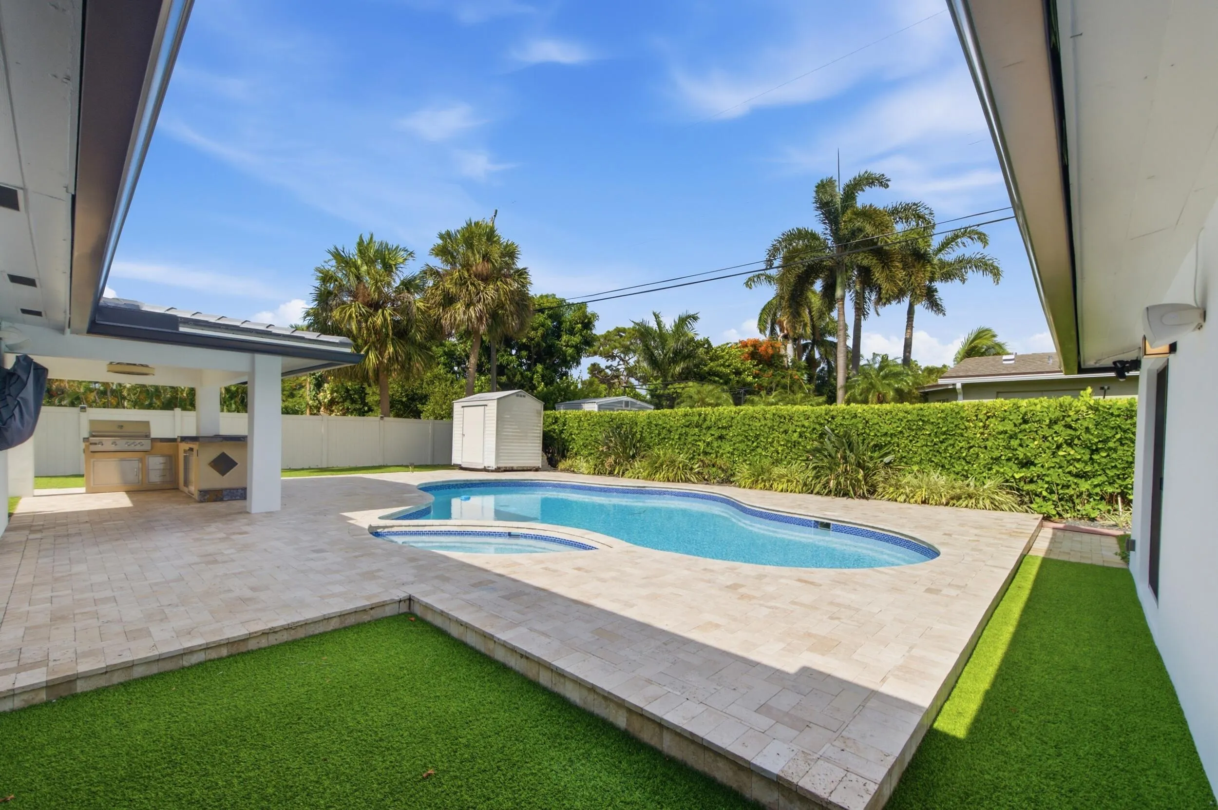 Resort-style backyard featuring a freeform swimming pool with integrated spa, expansive paver patio, covered outdoor kitchen with built-in grill, and artificial turf surrounds