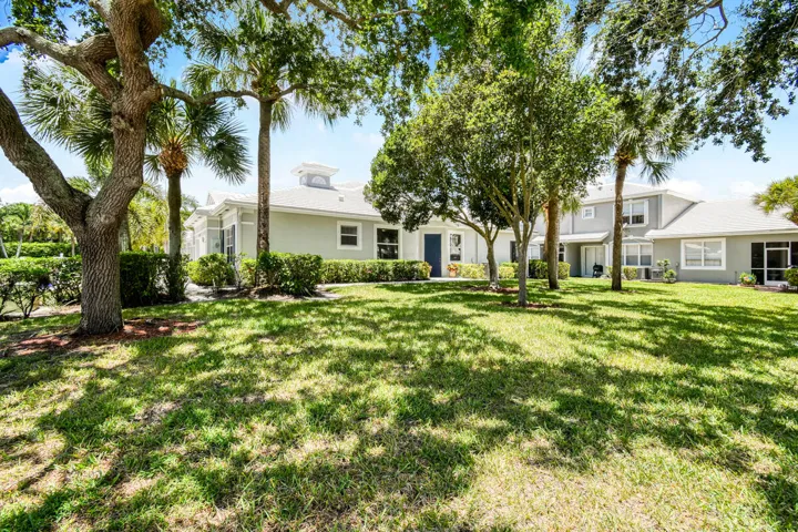 Expansive green lawn framed by mature trees and palm trees, leading to a multi-unit property with a light-colored exterior, multiple windows, and a distinctive cupola