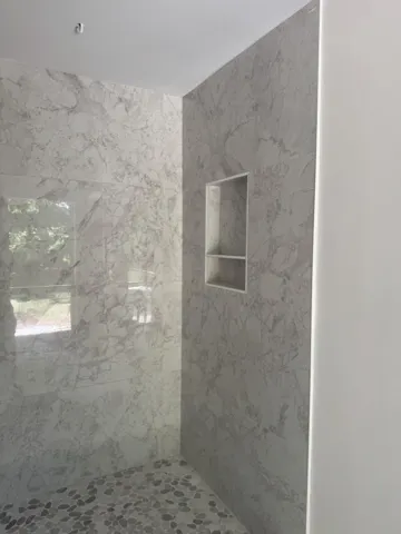 Shower enclosure featuring light-toned, veined tile walls, a built-in recessed niche, and a pebble-style floor