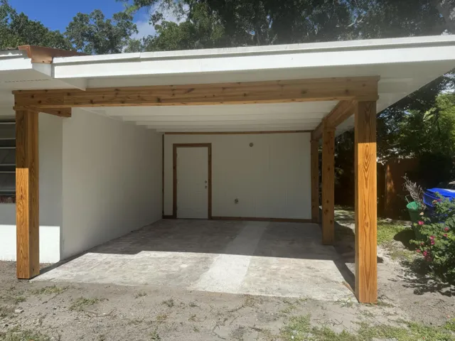 Covered outdoor patio area featuring a concrete slab, exposed wood support beams, and a white exterior wall with a service door