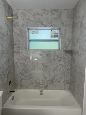 Bathtub with light gray veined wall tile surround, featuring a horizontal window and a built-in shelf
