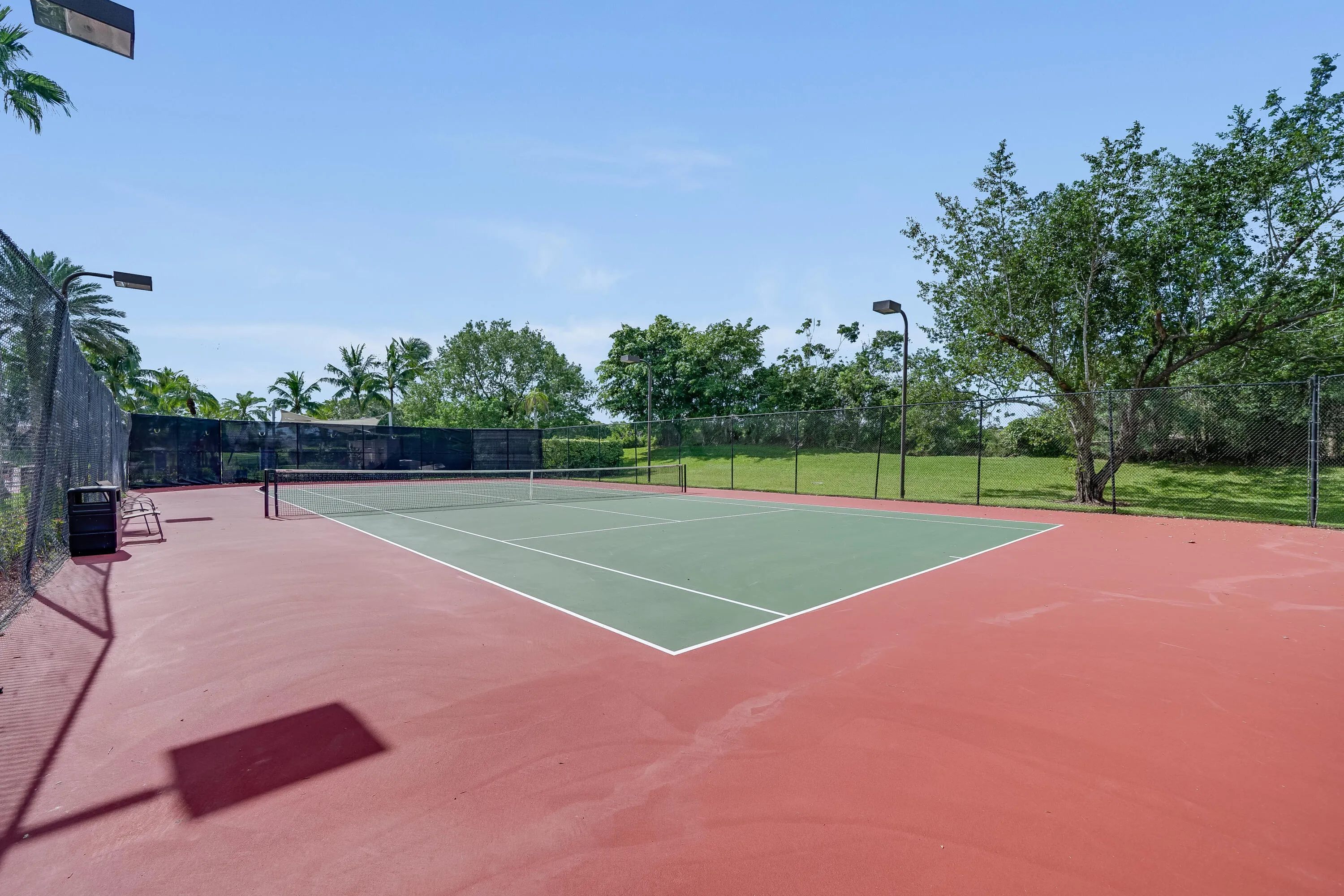 Regulation-size tennis court featuring a red and green surface, surrounding chain-link fencing, court lighting, and palm trees