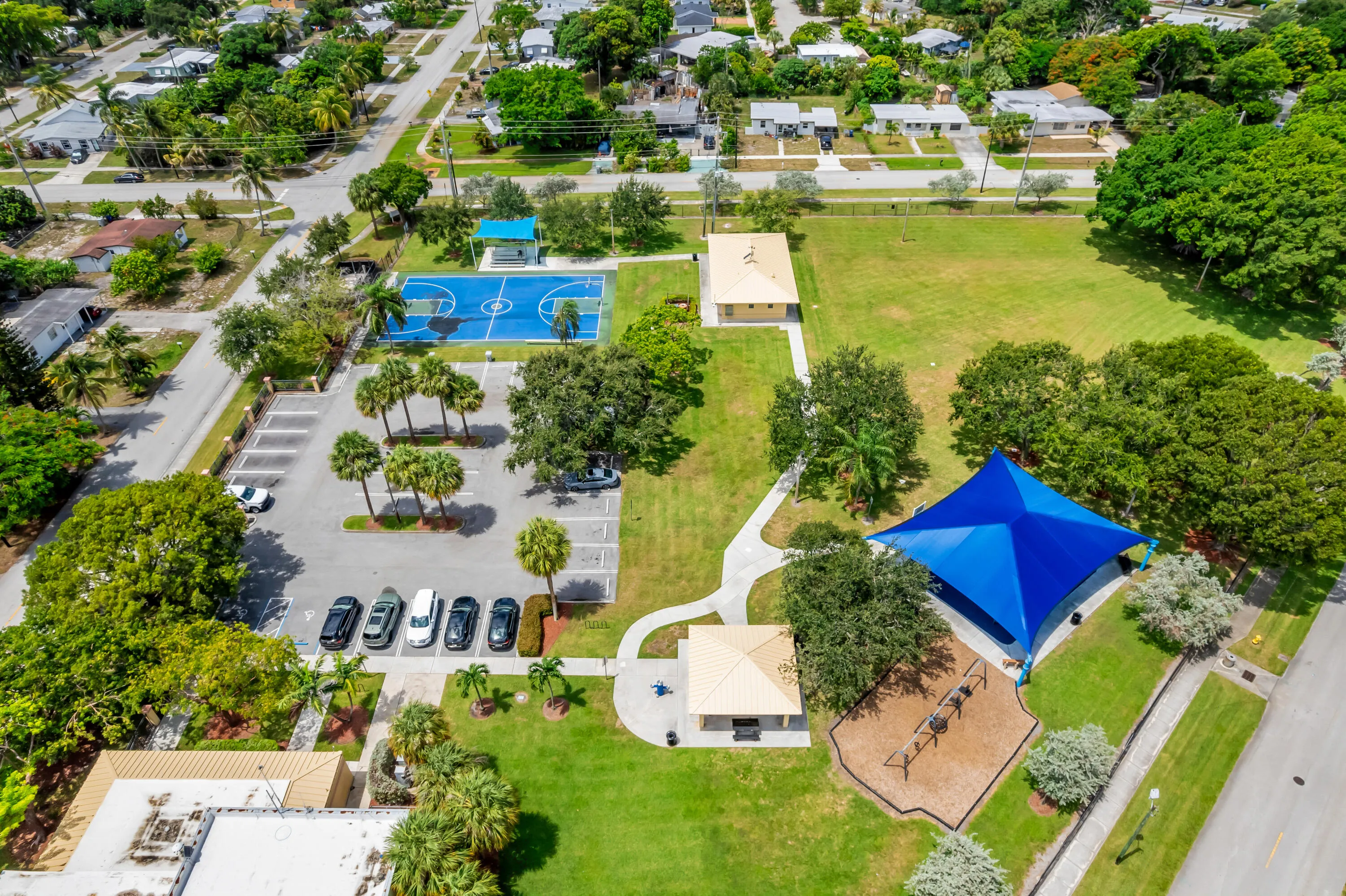 Expansive community park featuring a vibrant blue basketball court, shaded playground area, multiple paved walking paths, open green spaces, and ample parking
