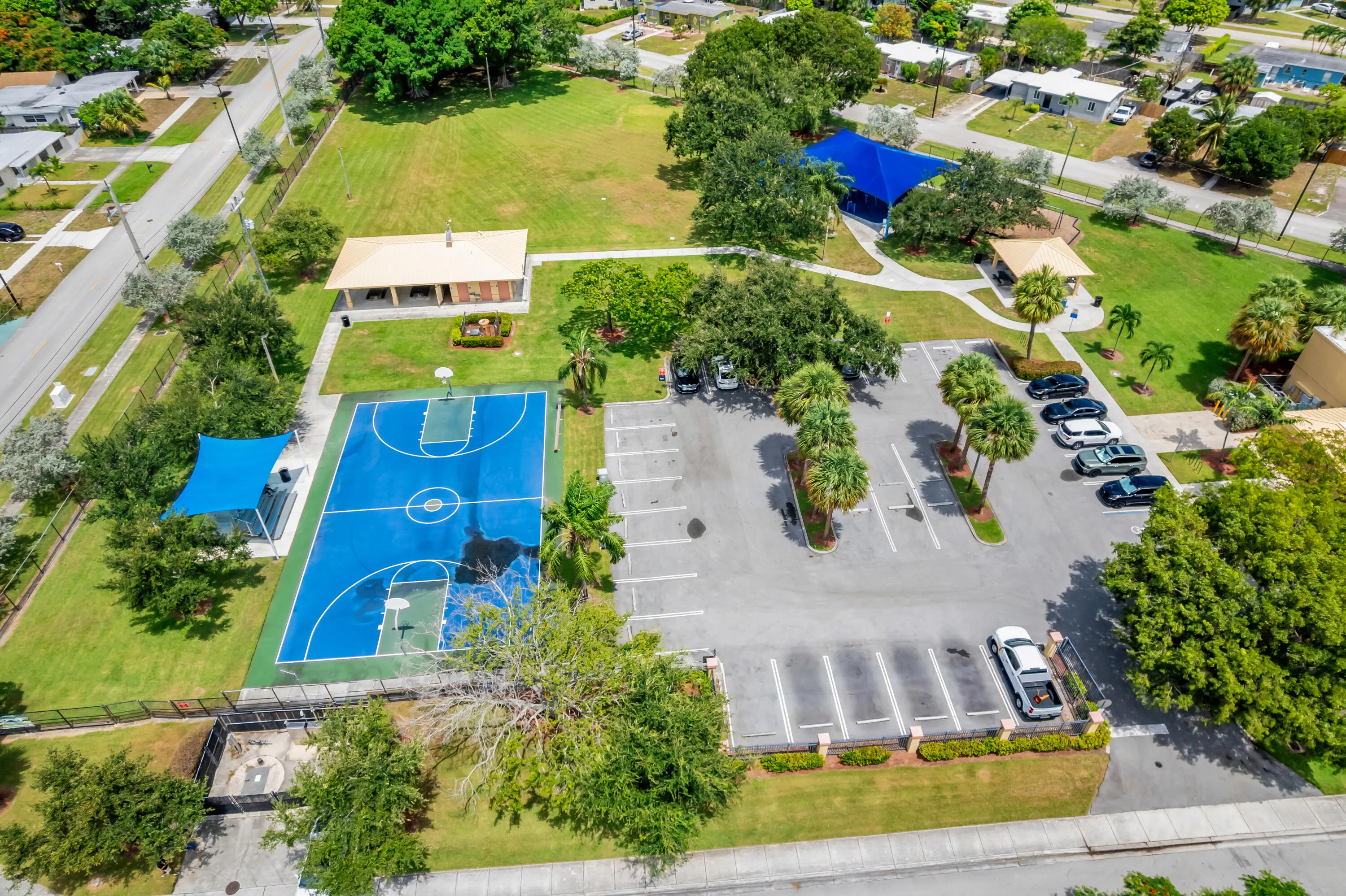 Recreational park grounds featuring a full-size basketball court, covered seating areas, and a paved parking lot with mature palm trees
