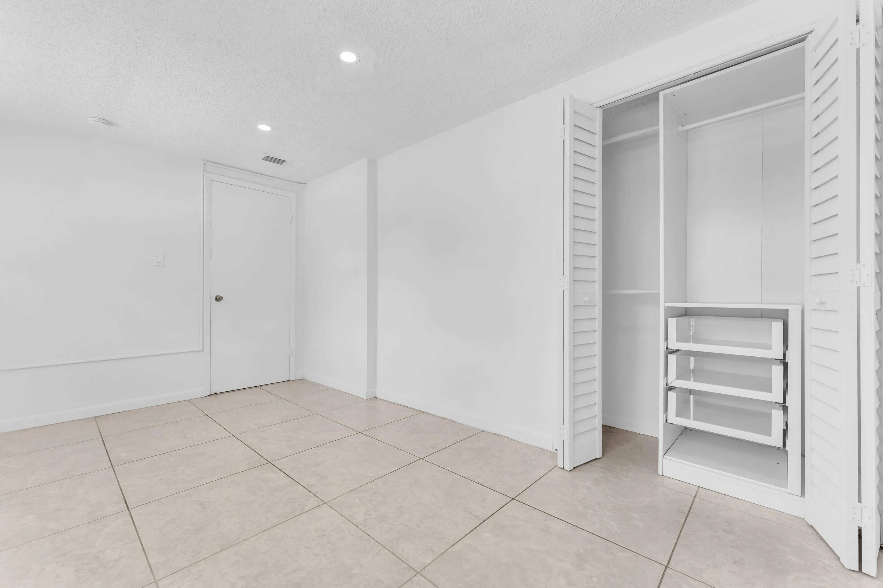 Bright room featuring neutral tile flooring, white walls, recessed lighting, and a closet with louvered bifold doors and built-in shelving