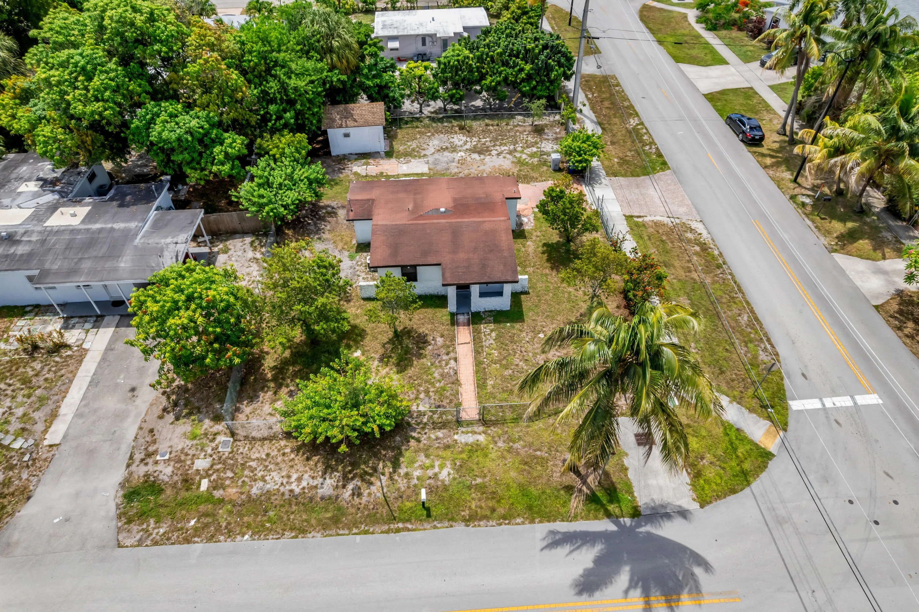 Aerial view showcasing a residential property with a brown-roofed structure, featuring a front yard with mature trees and a paved driveway