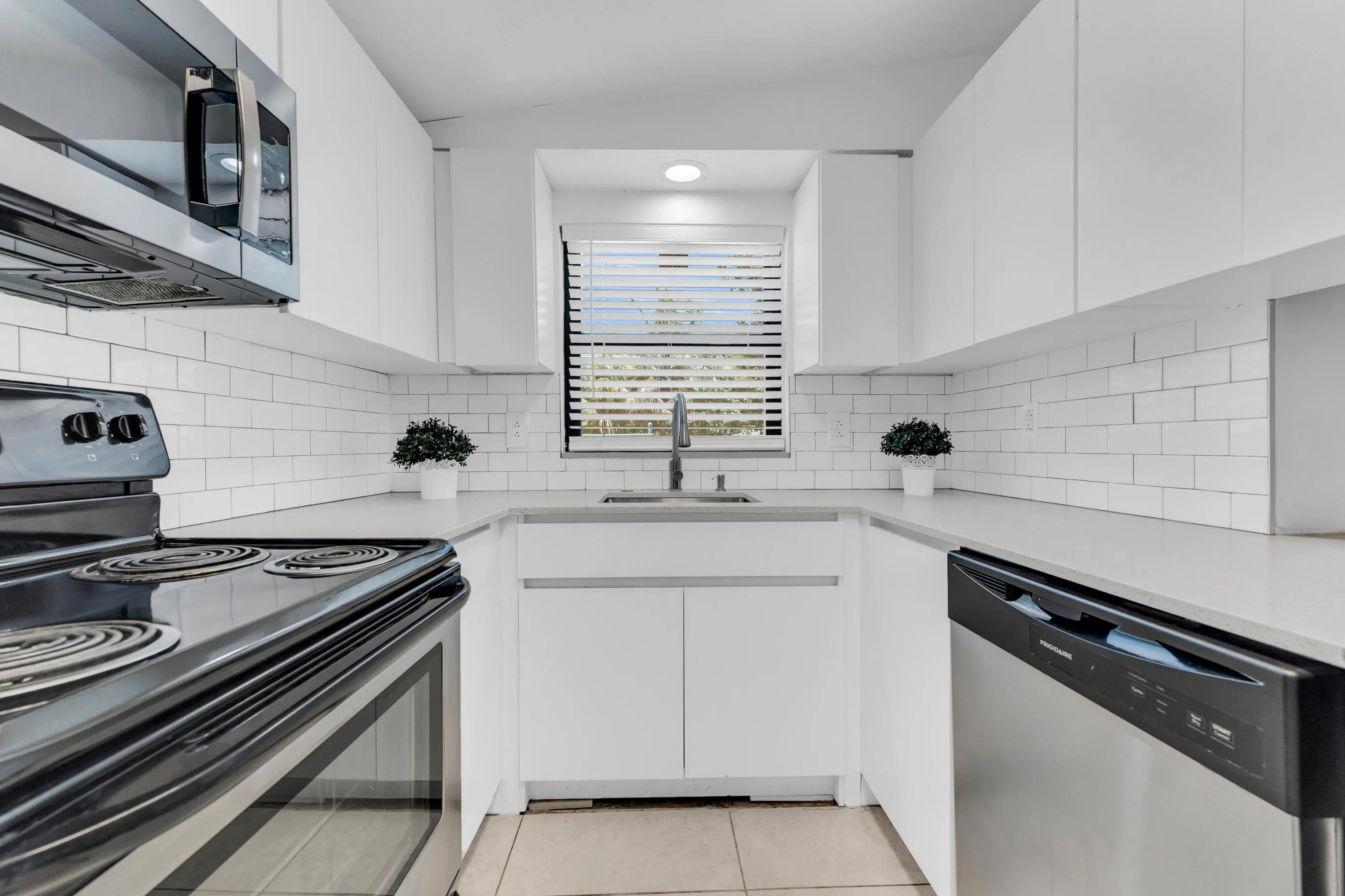 Modern kitchen featuring sleek white cabinetry, light-toned countertops, a white subway tile backsplash, stainless steel appliances, and a window with horizontal blinds