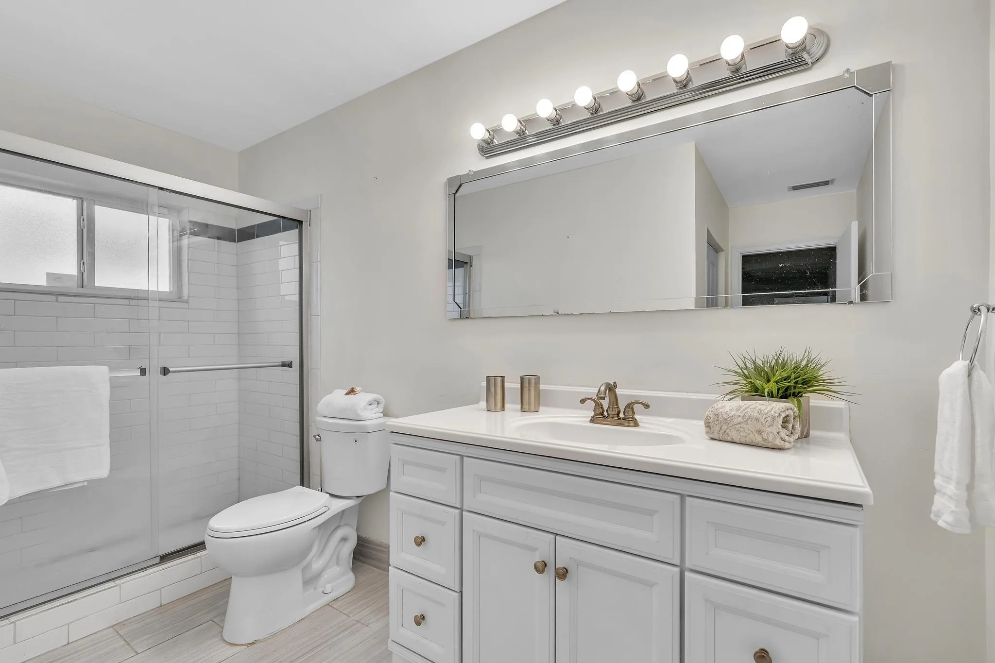 Bathroom featuring a sliding glass shower enclosure, white subway tile shower surround, a window within the shower, a single vanity with white cabinetry and a light-toned countertop, and a large wall-mounted mirror with a horizontal light fixture