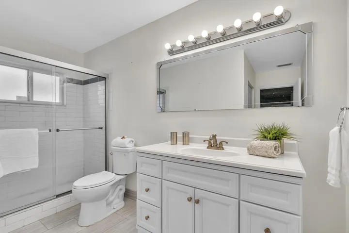 Bathroom featuring a sliding glass shower enclosure, white subway tile shower surround, a window within the shower, a single vanity with white cabinetry and a light-toned countertop, and a large wall-mounted mirror with a horizontal light fixture