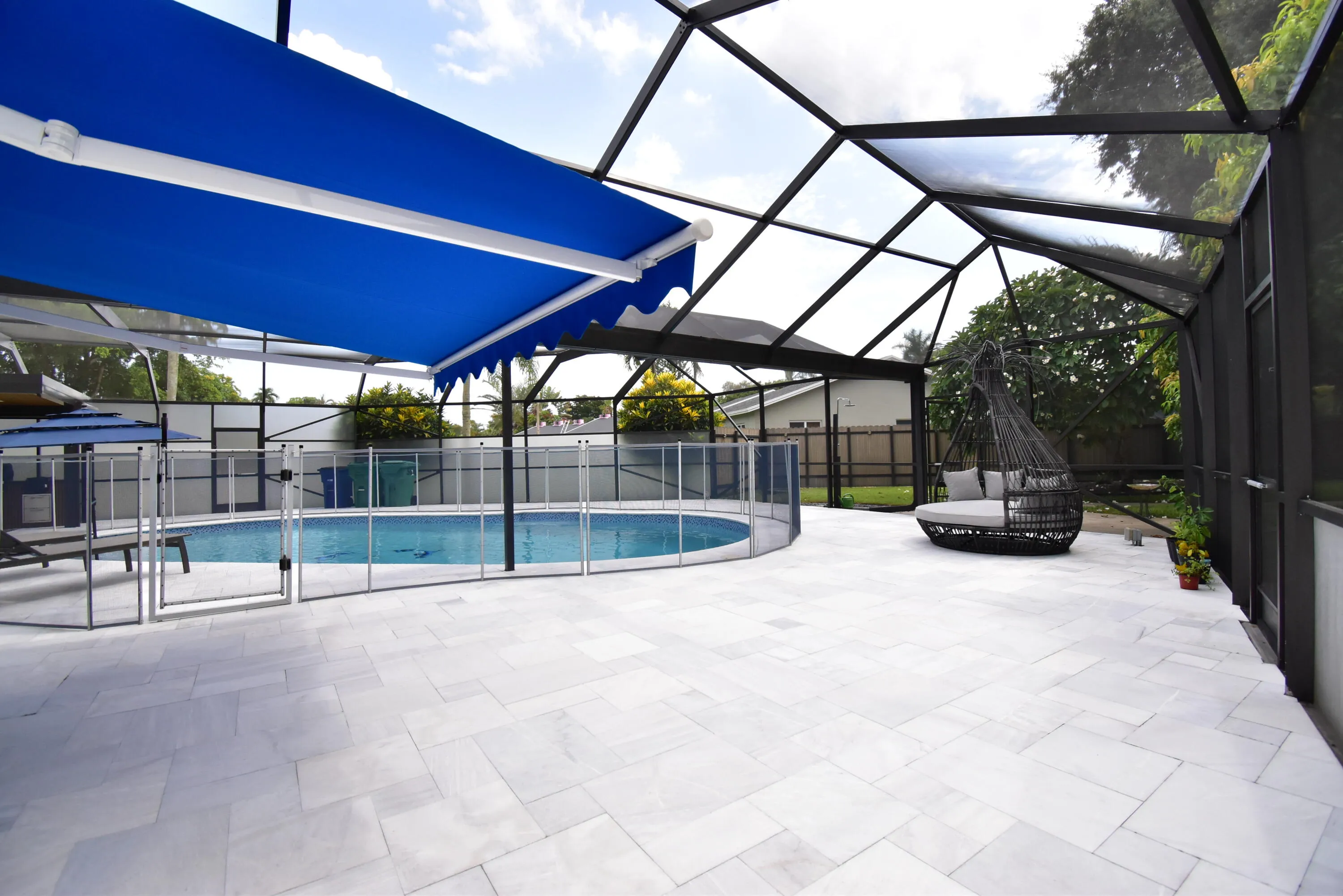 Spacious screened patio featuring an enclosed swimming pool, a blue retractable awning, a safety fence around the pool, and a light-toned paver deck