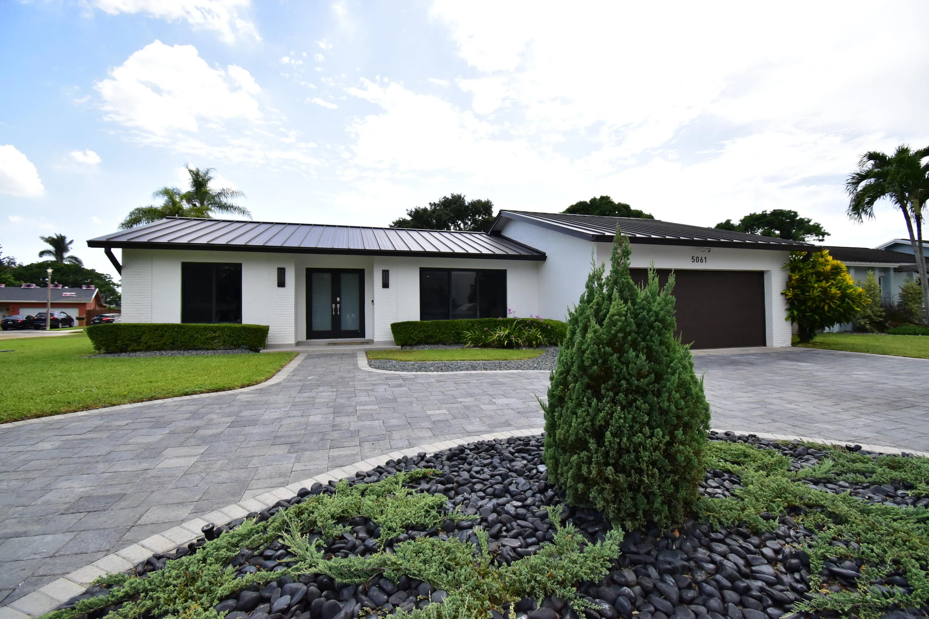 Contemporary white exterior featuring a dark metal roof, a paved circular driveway, a dark garage door, and a manicured front lawn with evergreen landscaping