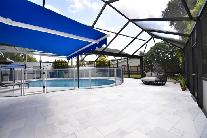 Spacious screened patio featuring an enclosed swimming pool, a blue retractable awning, a safety fence around the pool, and a light-toned paver deck