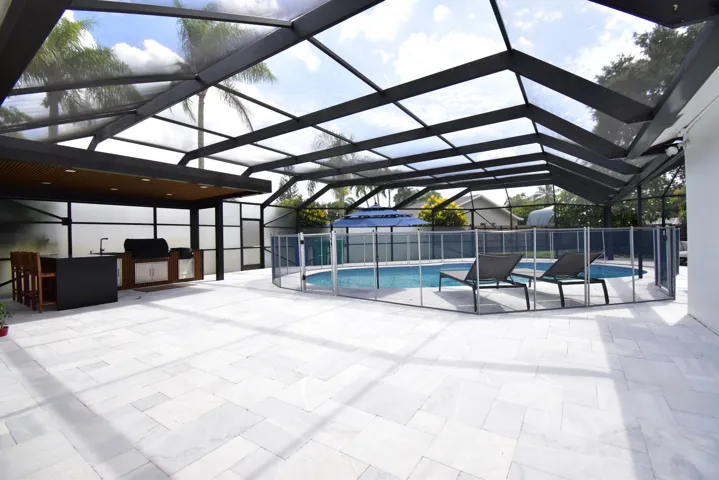 Expansive screened patio featuring light-toned paver flooring, a built-in outdoor kitchen with a sink and grill, and a fenced swimming pool