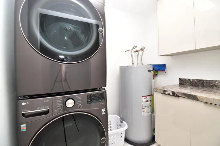 Laundry area featuring a stacked front-load washer and dryer, a water heater, a stone-finish countertop, and upper and lower cabinetry with integrated pulls