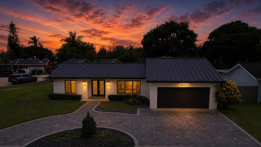 Contemporary exterior featuring a dark standing seam metal roof, white painted brick facade, a two-car garage, and a paver driveway