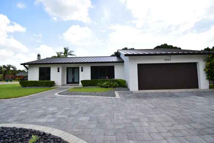 Contemporary white brick exterior featuring a dark metal roof, a paved driveway, manicured landscaping, and a two-car garage