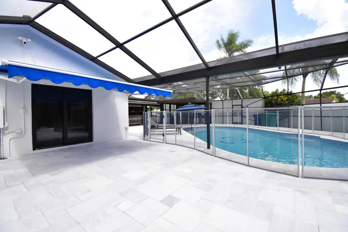 Screened-in pool area featuring a rectangular swimming pool with a mosaic tile interior, a safety fence, and an expansive light-toned paver deck