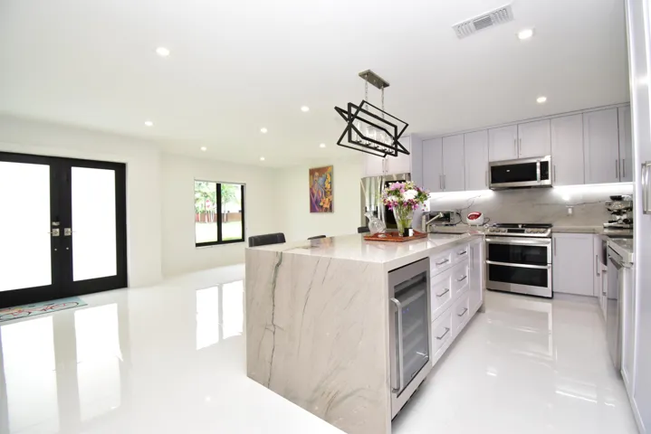 Modern kitchen featuring a waterfall island with integrated wine refrigerator, stainless steel appliances, light cabinetry, recessed lighting, and white tile flooring