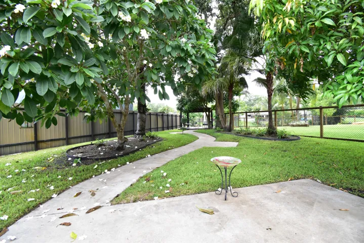Expansive backyard featuring a concrete patio, winding concrete pathway, mature trees, and a chain-link fence