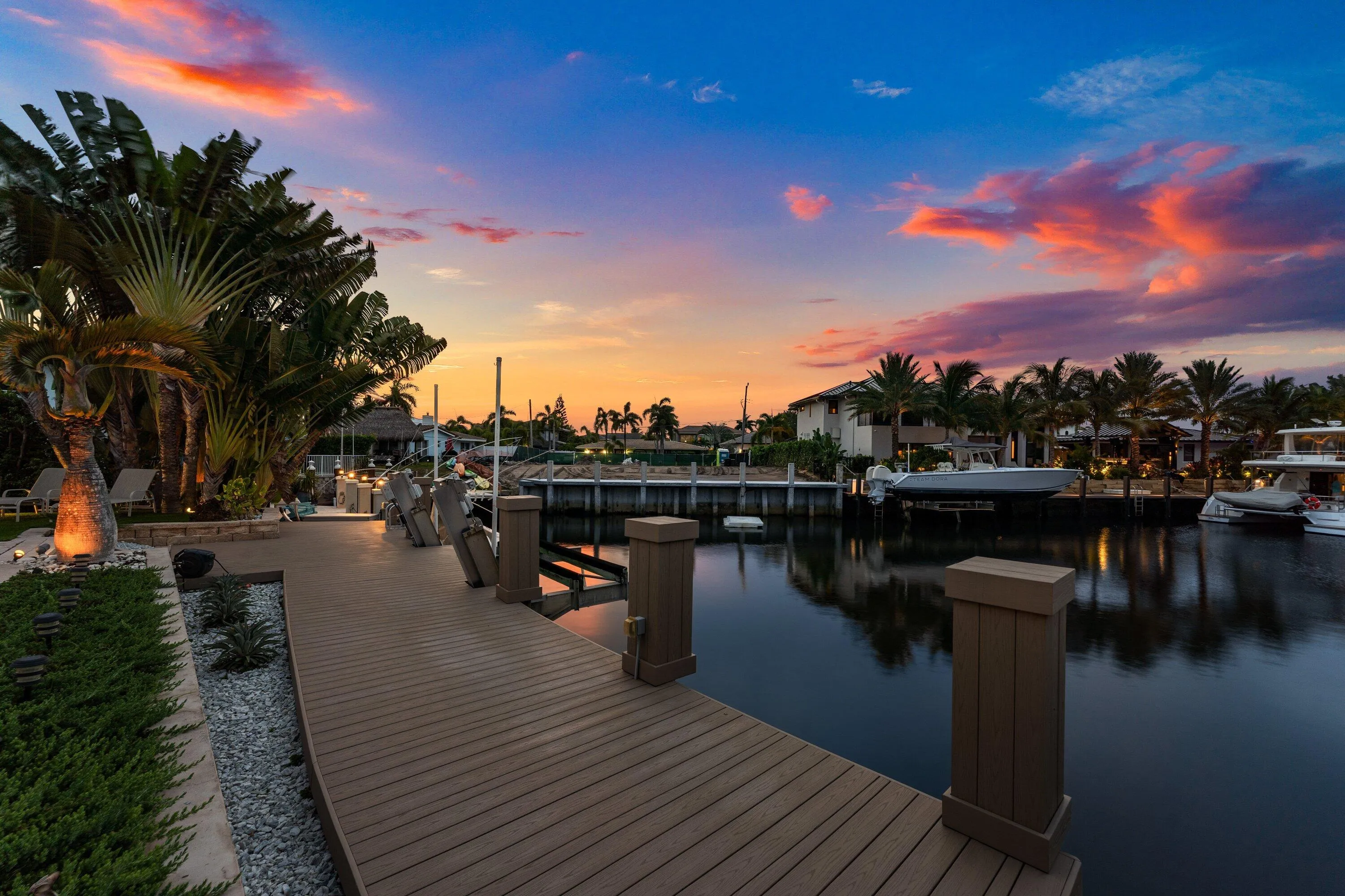 Waterfront property featuring an expansive composite deck, mature palm trees, integrated landscape lighting, and a private boat slip