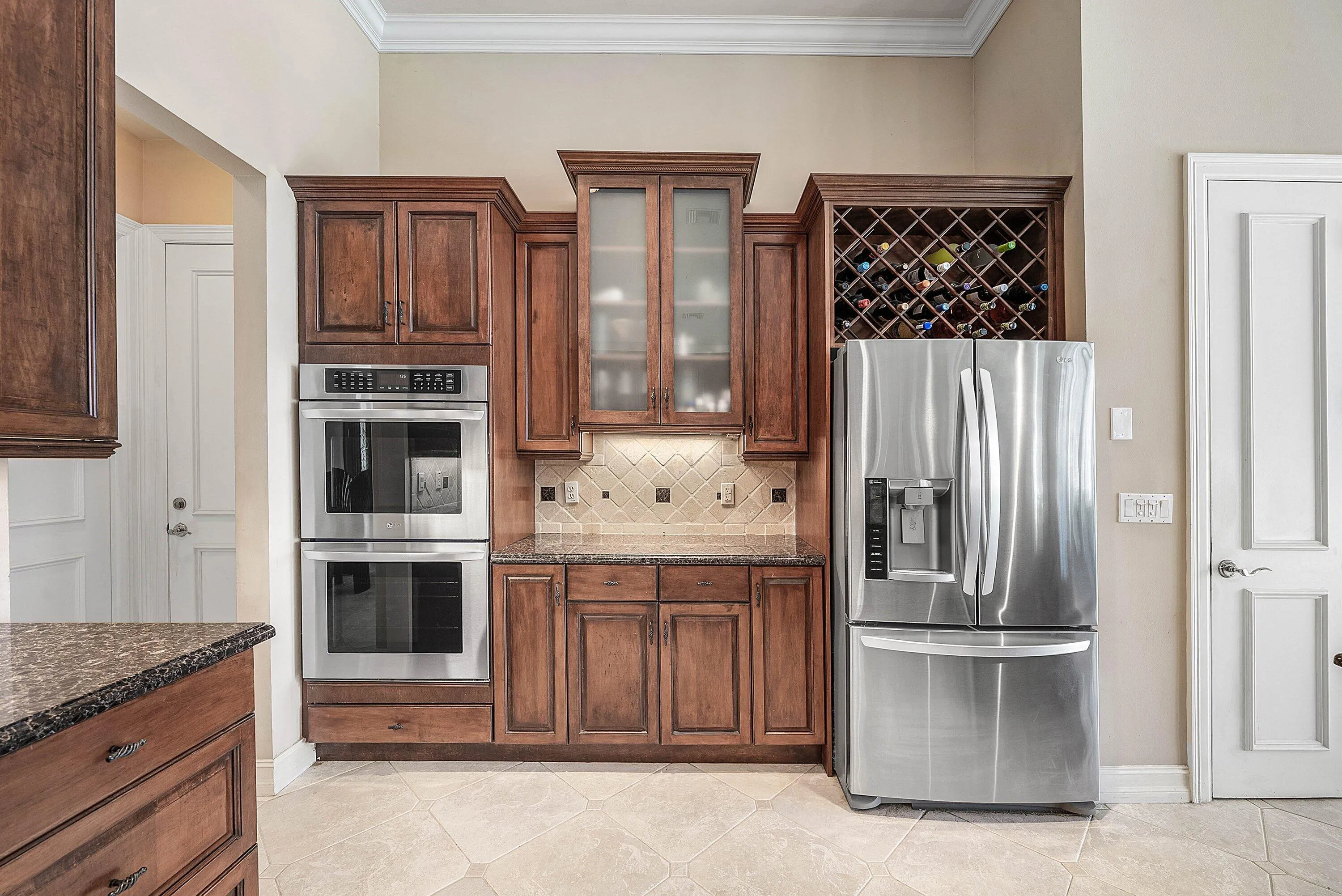 Kitchen appliance wall featuring dark wood-finish cabinetry, stainless steel double ovens, a built-in wine rack, a French door refrigerator, and a tiled backsplash