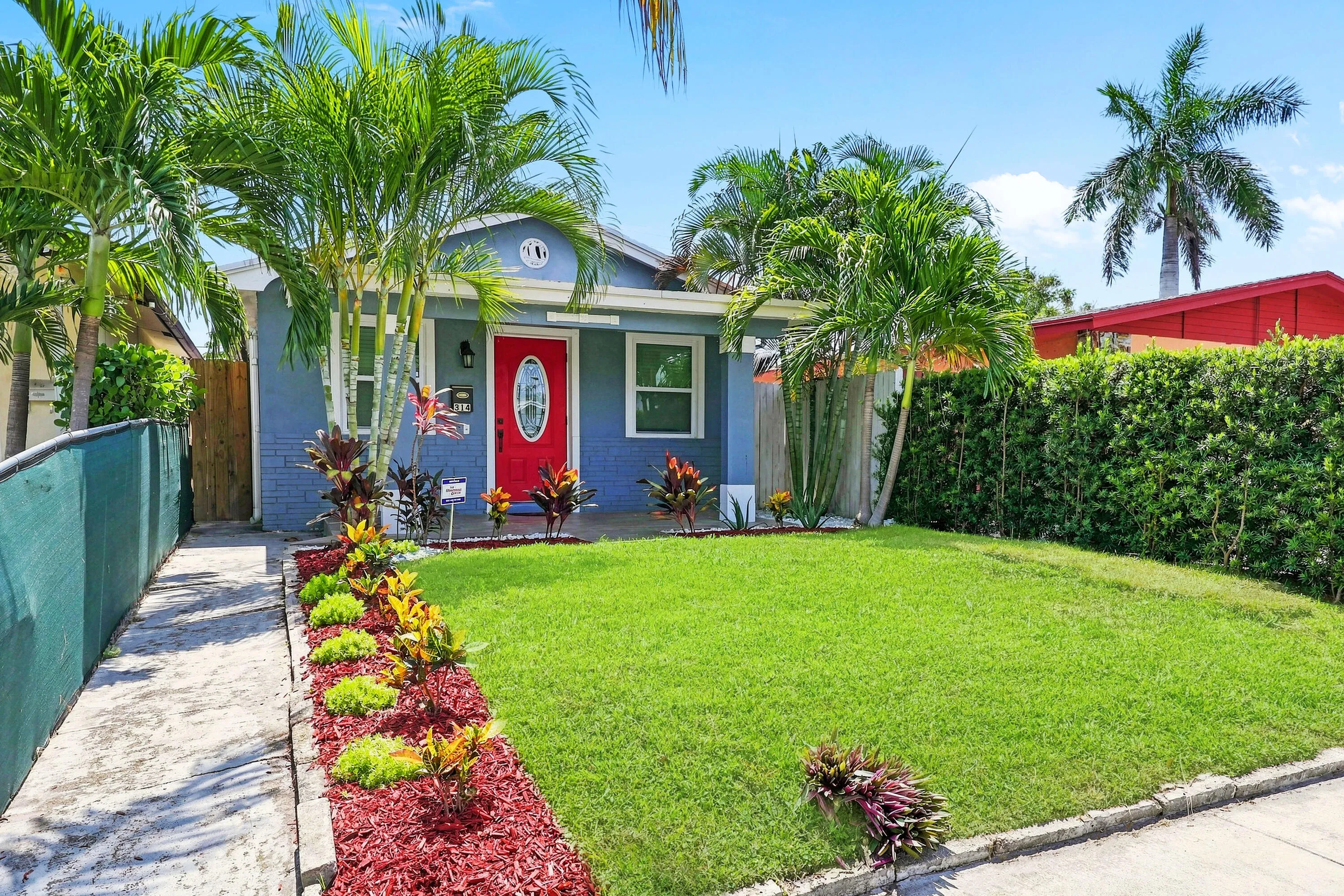 Charming blue exterior featuring a vibrant red front door, manicured lawn, tropical landscaping with palm trees, and a concrete walkway