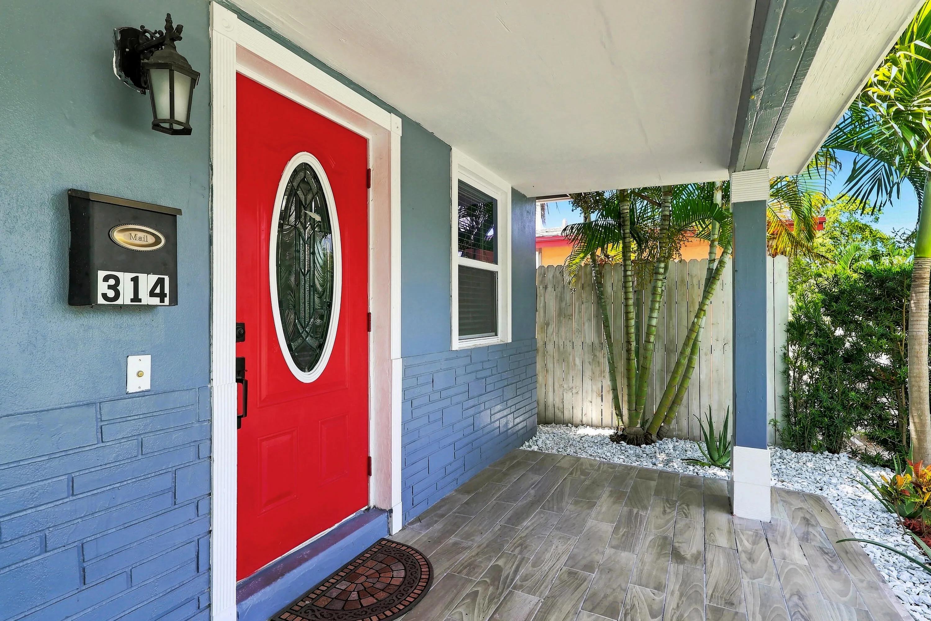 Covered front porch featuring wood-finish flooring, a vibrant red entry door with an oval stained-glass insert, a wall-mounted exterior light fixture, and a decorative privacy fence