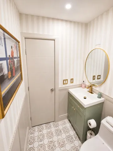 Powder room featuring patterned floor tiles, striped wallpaper, a green vanity with integrated sink, and a gold-framed circular mirror