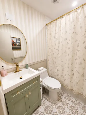 Bathroom featuring a sage green vanity with gold hardware, a circular gold-framed mirror, a modern white toilet, patterned floor tiling, and striped wallpaper