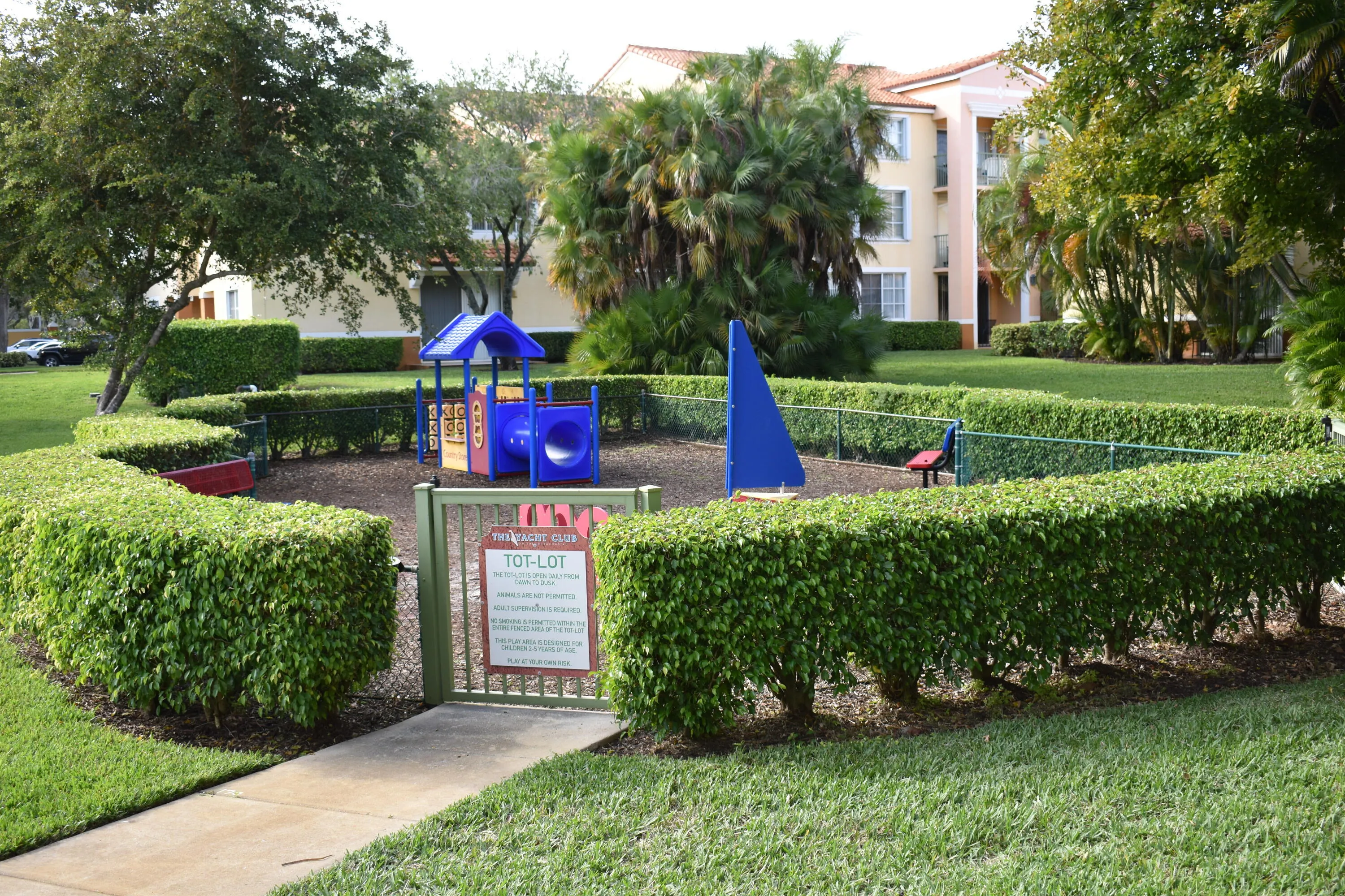 Community tot-lot featuring a play structure with slide, fenced perimeter with hedge landscaping, and a gated entry