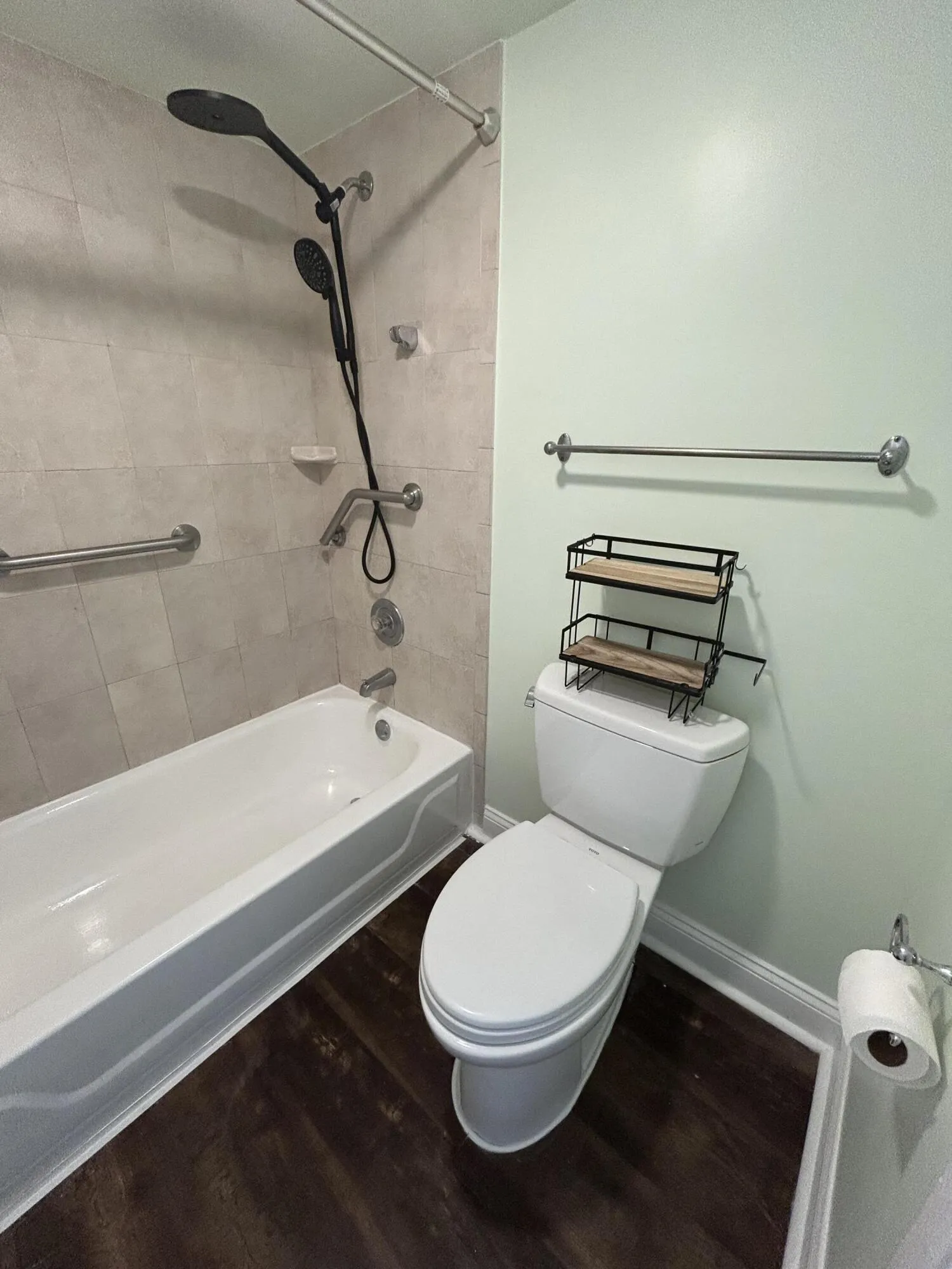 Bathroom featuring a white bathtub with a tiled surround, grab bars, a dual shower head system, and dark wood-finish flooring