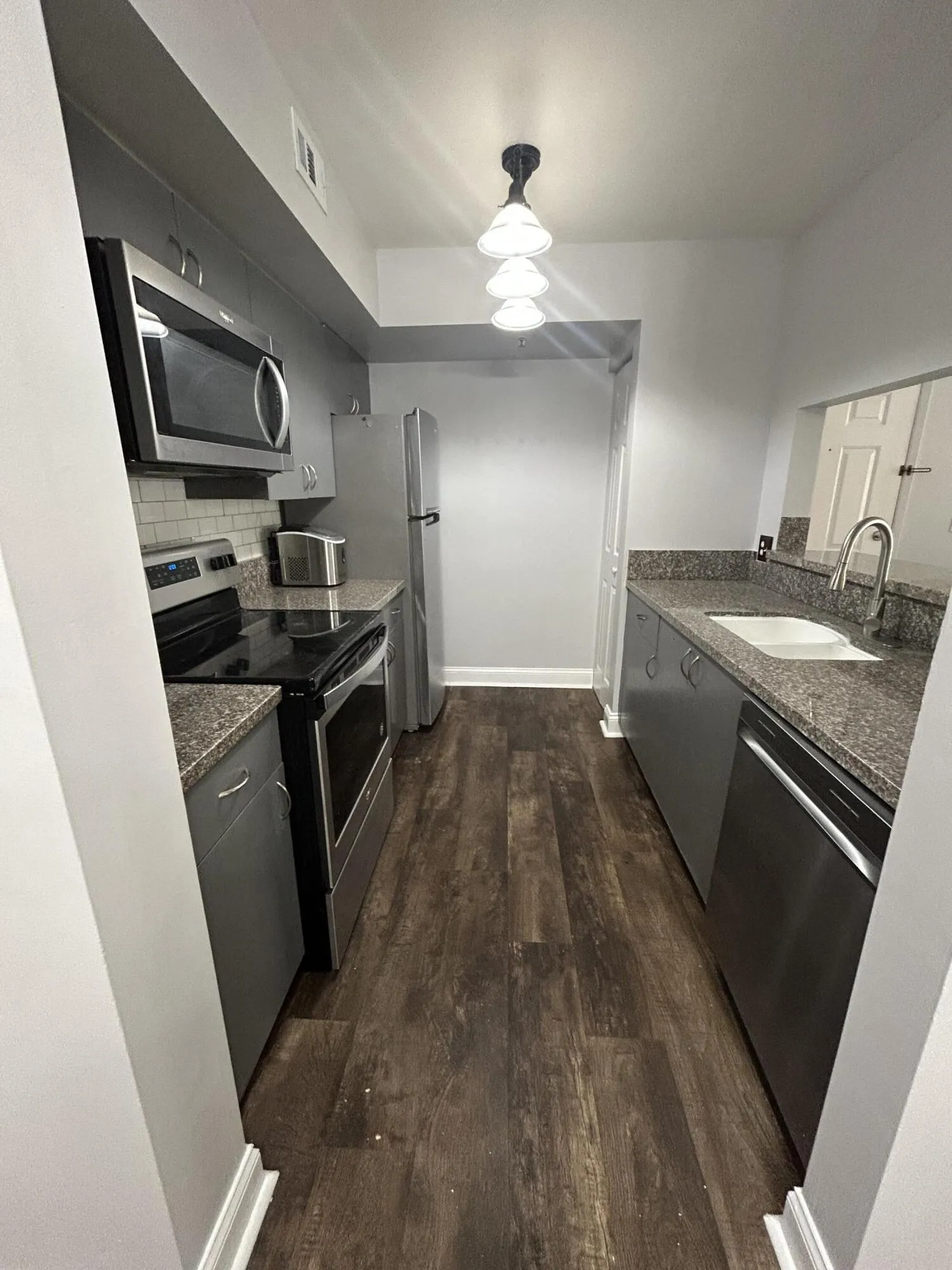 Galley kitchen featuring wood-finish flooring, stainless steel appliances, grey cabinetry, granite-style countertops, and a tiled backsplash