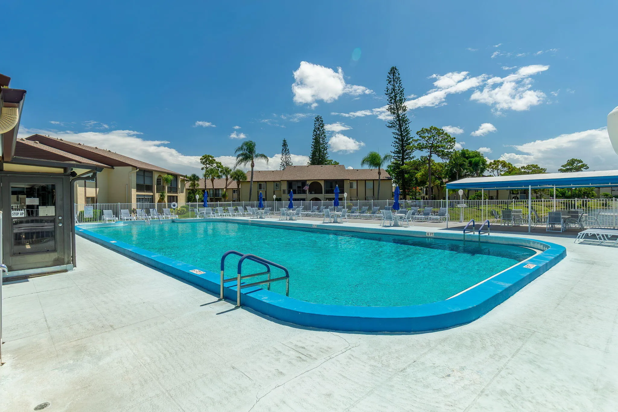 Community pool featuring a large blue-tiled swimming area, surrounding concrete decking, multiple lounge chairs, and a covered seating area